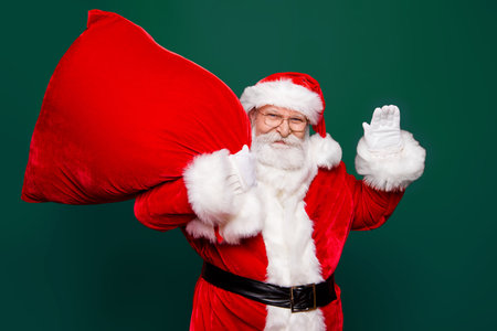 Welcome to festive event party! Careless carefree grandfather stylish Santa in headwear spectacles white beard hold bag on shoulder look at camera waving goodbye or hello isolated on red backgroundの写真素材