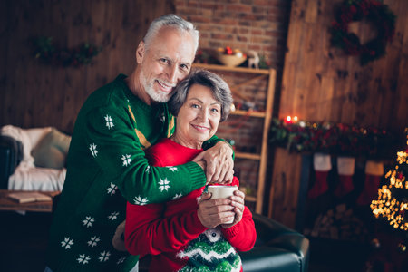 Photo of positive cheerful couple hugs old age seniors staying home wear comfortable sweaters hold cup sweat drink hot cacao indoors xmas atmosphereの写真素材