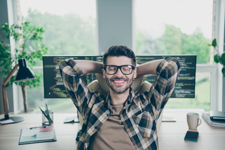 Photo of smiling positive programmer wear spectacles relaxing after working device hands behind head...