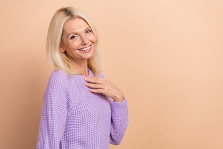 Portrait of charming retired woman with straight hairstyle dressed violet sweater hold palm on chest isolated on beige color backgroundの写真素材
