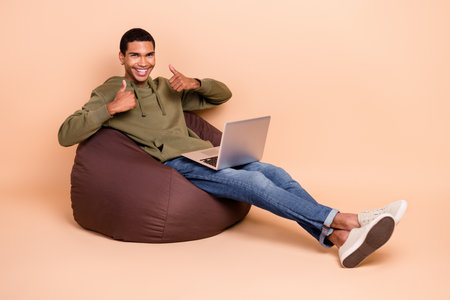 Photo of joyful man sitting comfort beanbag thumbs up recommend nice modern device sale empty space isolated on beige color backgroundの写真素材