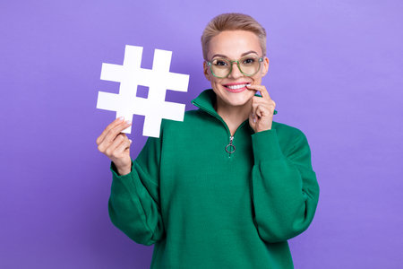Photo of lovely smile short blonde hair girl bite finger dreaming million followers hold popular hashtag symbol isolated on violet color backgroundの写真素材