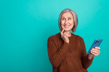 Photo of positive aged lady finger touch chin hold telephone look empty space isolated on teal color...