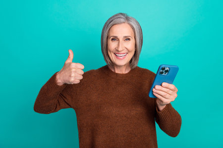 Photo of positive charming grandmother hold telephone demonstrate thumb up isolated on teal color...