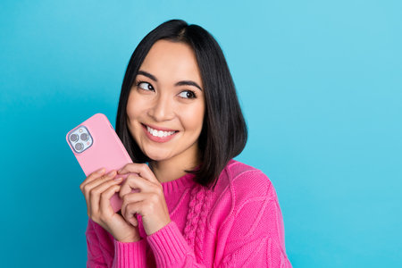 Portrait of cute cheerful thai girl hold telephone toothy smile look interested empty space isolated on blue color backgroundの写真素材
