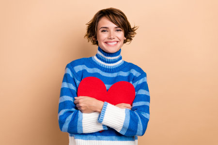 Photo of sweet shiny lady wear blue pullover smiling embracing red heart isolated beige color backgroundの写真素材