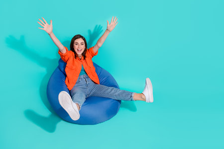 Full length photo of carefree funny lady wear orange shirt sitting bean bag rising arms empty space...