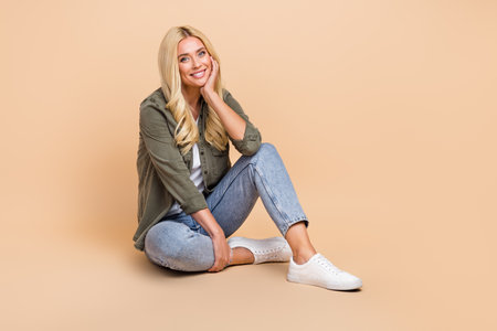 Full length photo of cheerful cute lady hand face wear khaki clothes sitting near empty space banner isolated on beige color backgroundの写真素材