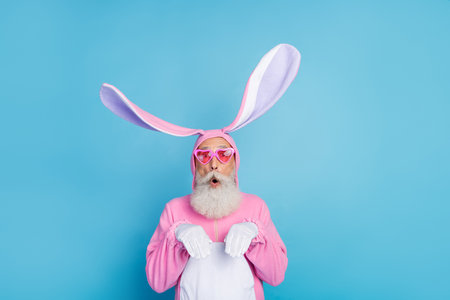 Photo of excited fooling man pensioner dressed pink rabbit nightwear heart eyewear rising arms hands isolated blue color backgroundの写真素材