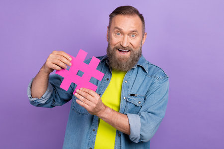 Portrait of cheerful aged person toothy smile arms hold paper hashtag symbol isolated on purple...