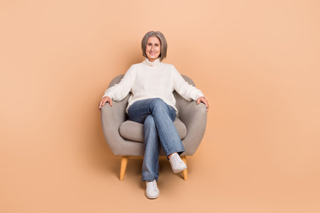Full length photo of cute charming woman wear knitted pullover sititng comfortable chair isolated beige color backgroundの写真素材