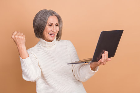 Photo of lucky pretty woman wear knitted pullover rising fist winning egame modern gadget isolated beige color backgroundの写真素材