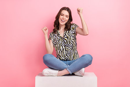 Full body photo of overjoyed crazy lady sitting platform raise fists up celebrate discount proposition shopping isolated on pink color backgroundの写真素材