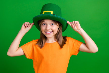 Photo of charming positive schoolchild toothy smile hands touch hat isolated on green color backgroundの写真素材