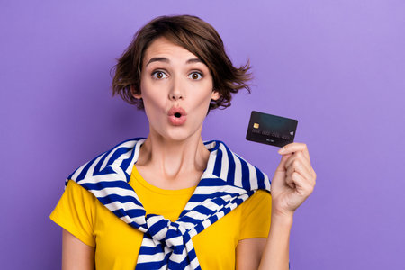 Photo of positive excited woman wear tied sweater big eyes showing credit card empty space isolated purple color backgroundの写真素材