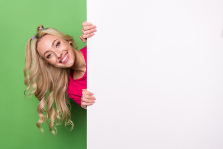 Photo of gorgeous funky lady toothy smile peeking behind empty space placard isolated on green color backgroundの写真素材