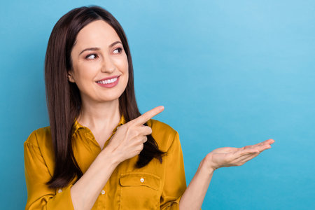 Portrait of beautiful promoter young brunette woman wear formal yellow shirt hold palm finger point look mockup isolated on blue color backgroundの写真素材