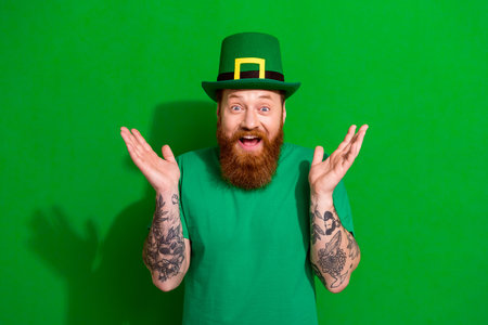 Photo of funny impressed guy dressed t-shirt cap rising arms celebrating clover day isolated green color backgroundの写真素材