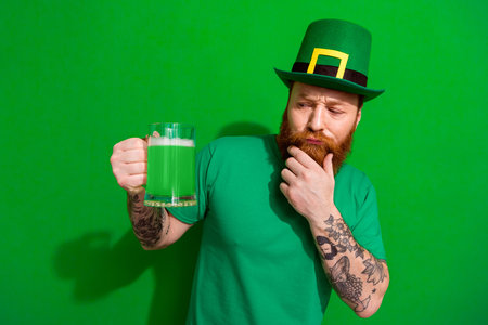 Photo of doubtful unsure guy dressed t-shirt cap celebrating clover day looking ale beer arm chin isolated green color backgroundの写真素材