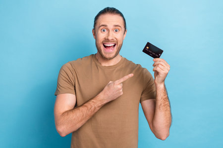 Photo of entrepreneur man wear t-shirt direct finger surprised demonstrate plastic credit card wireless nfc payment isolated on blue color backgroundの写真素材