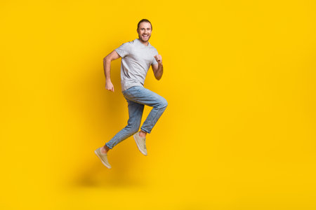Full length photo of excited funky guy dressed grey t-shirt jumping high running fast empty space isolated yellow color backgroundの写真素材