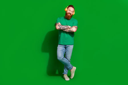 Full length photo of positive cool man wear t-shirt headphones arms folded isolated green color backgroundの写真素材