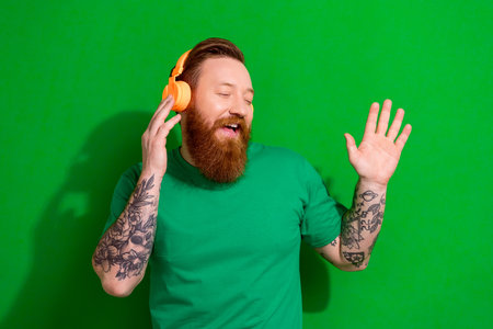 Photo of handsome cheerful guy ginger beard dressed stylish t-shirt headphones singing song at home isolated on green color backgroundの写真素材