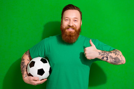 Portrait of cheerful positive man with red beard wear trendy t-shirt hold football ball show like isolated on green color backgroundの写真素材