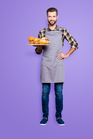 Full size body portrait of positive attractive baker in jeans, shoes, shirt, apron with stubble having tray with bakery products, looking at camera, isolated on grey backgroundの写真素材