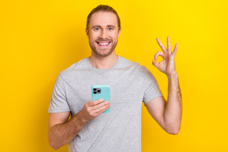 Photo of cheerful confident guy dressed grey t-shirt typing device showing okey isolated yellow color backgroundの写真素材
