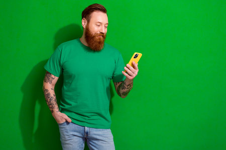 Photo of masculine irish guy with ginger beard dressed stylish t-shirt look at phone typing post isolated on green color backgroundの写真素材