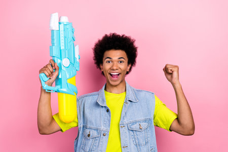 Photo of cheerful delighted person hold water gun raise fist good mood isolated on pink color backgroundの写真素材