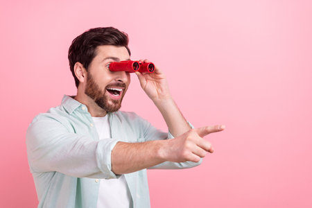 Photo portrait of handsome young guy hold binoculars excited point finger dressed stylish blue clothes isolated on pink color backgroundの写真素材
