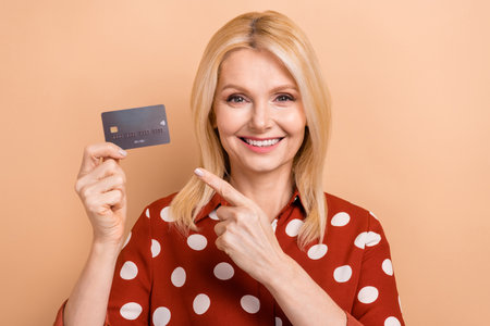 Portrait of positive good mood woman with blond hair dressed red shirt directing at credit card isolated on beige color backgroundの写真素材