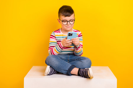 Photo of cute charming boy wear trendy clothes sit platform cube use modern technology device isolated on yellow color backgroundの写真素材