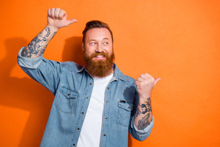 Photo of cool cheerful man wear jeans shirt pointing two fingers looking empty space isolated orange color backgroundの写真素材
