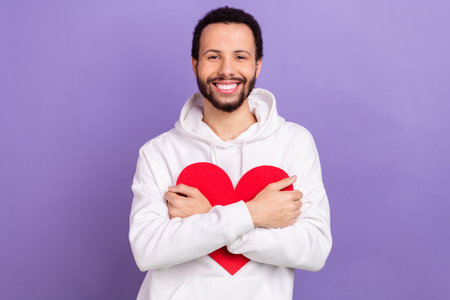 Photo of cheerful candid man beaming smile arms hold cuddle paper red heart isolated on violet color backgroundの写真素材