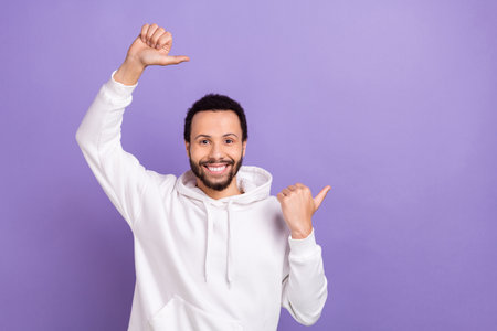 Photo of friendly man with curly hairstyle dressed white hoodie directing at offer empty space isolated on purple color backgroundの写真素材