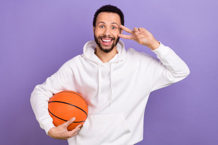 Photo of positive nice guy arm hold basketball demonstrate v-sign near eye isolated on violet color backgroundの写真素材