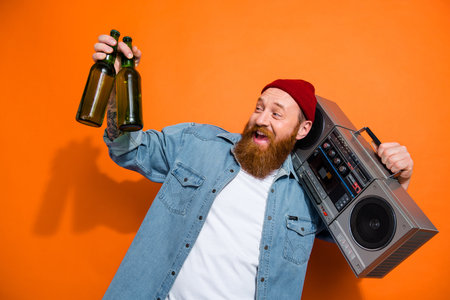 Photo of careless funny guy red hair beard wear beanie stylish cap denim jacket hold boombox drink glass bottle isolated on orange color backgroundの写真素材