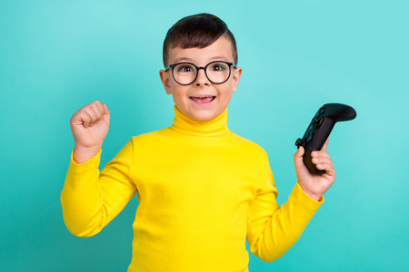 Photo of positive cheerful boy playing video game raise fists up winning isolated cyan color...