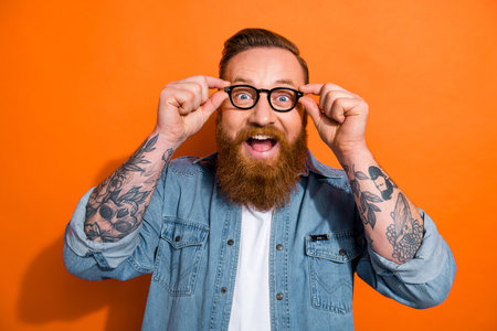 Photo of impressed funny man wear jeans shirt arms spectacles open mouth isolated orange color backgroundの写真素材
