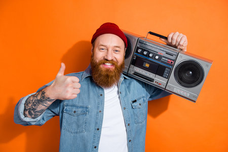 Photo of irish funny redhair guy thumb up dj music lover hold boom box retro party bearded tattooed person isolated on orange color backgroundの写真素材
