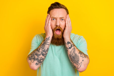 Photo of young irish handsome guy red hair touch cheeks shocked green t-shirt scared how bad his business goes isolated on yellow color backgroundの写真素材