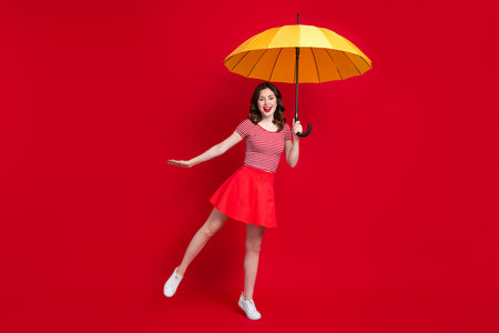 Full size portrait of carefree cheerful girl hand hold umbrella stand one leg isolated on red color backgroundの写真素材