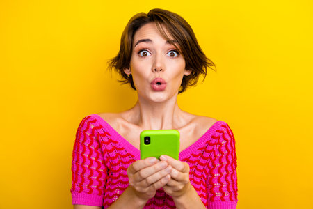 Portrait of astonished excited woman short haircut wear pink knit top read notification on smartphone isolated on yellow color backgroundの写真素材