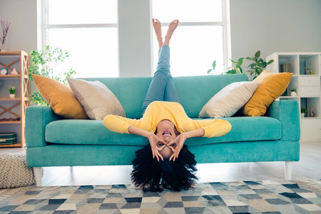 Full length portrait of cheerful careless person fingers hands make binoculars eye show tongue out upside down sofa house indoorsの写真素材