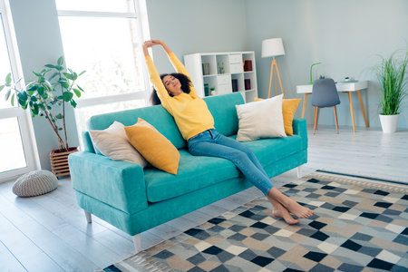Portrait of optimistic peaceful satisfied girl with wavy hairstyle dressed yellow sweatshirt stretching on sofa in morning indoors roomの写真素材