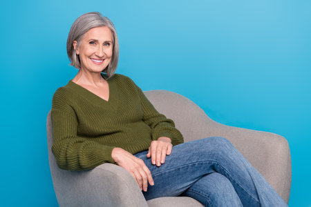 Photo of mature age woman grey hair wear khaki sweater sit grey armchair comfort room for psychotherapy isolated on blue color backgroundの写真素材