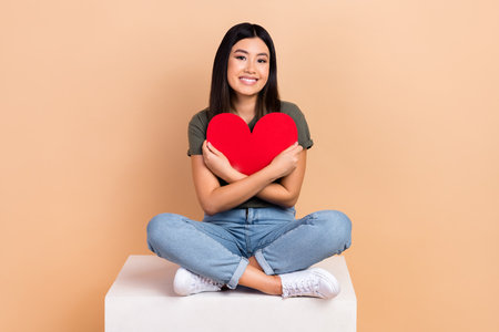 Full length photo of korean young lady sitting platform embracing red paper heart symbol feelings isolated on beige color backgroundの写真素材